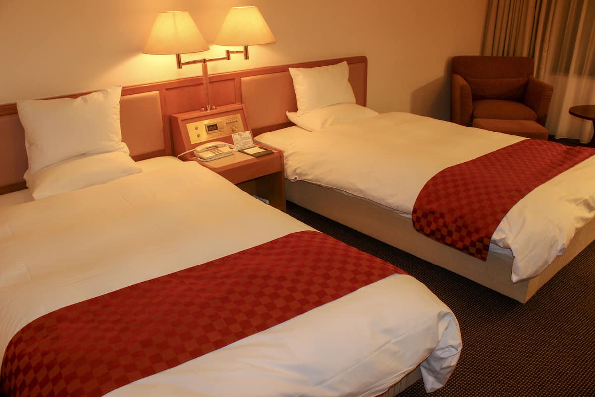 Habitación de hotel con dos camas, cada una con ropa blanca y una manta roja de cuadros. Hay dos lámparas de noche, una mesita de noche con un teléfono y un bloc de notas, y un sillón marrón.