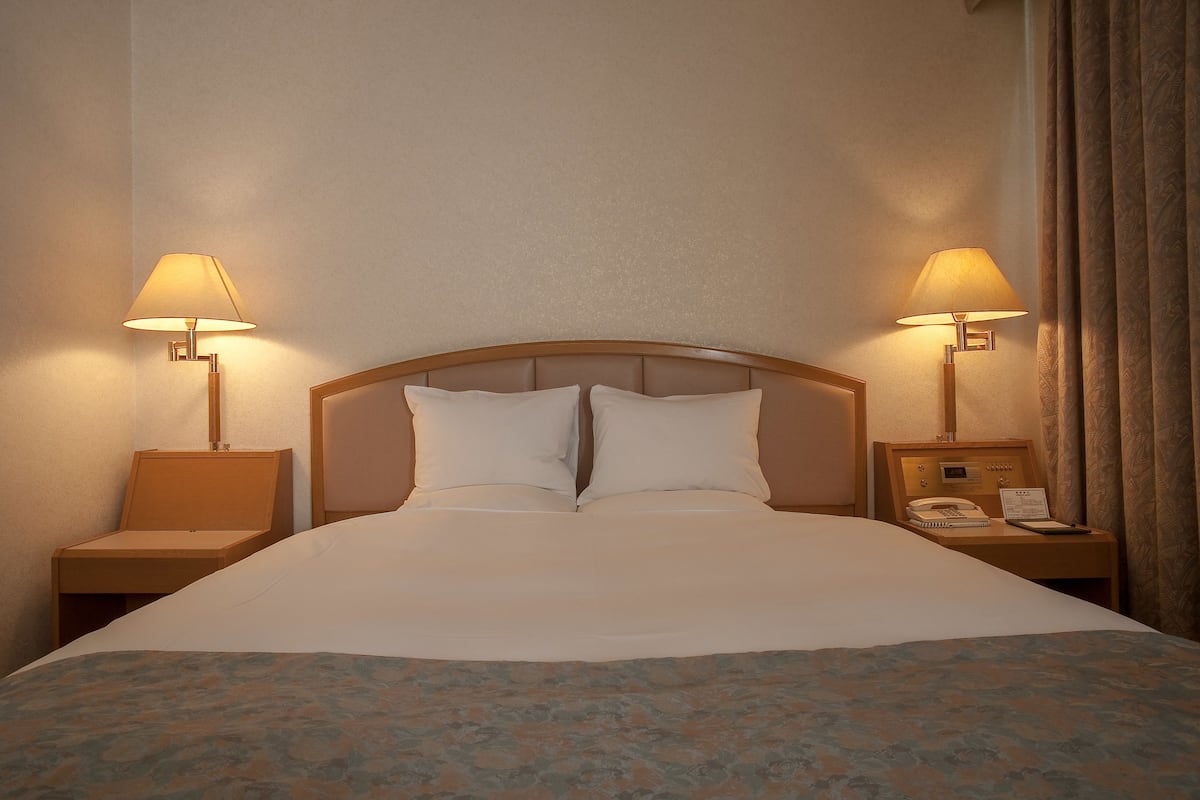 Habitación de hotel con una cama grande, dos lámparas de noche, una mesita de noche con un teléfono y una libreta, y una ventana con cortinas.