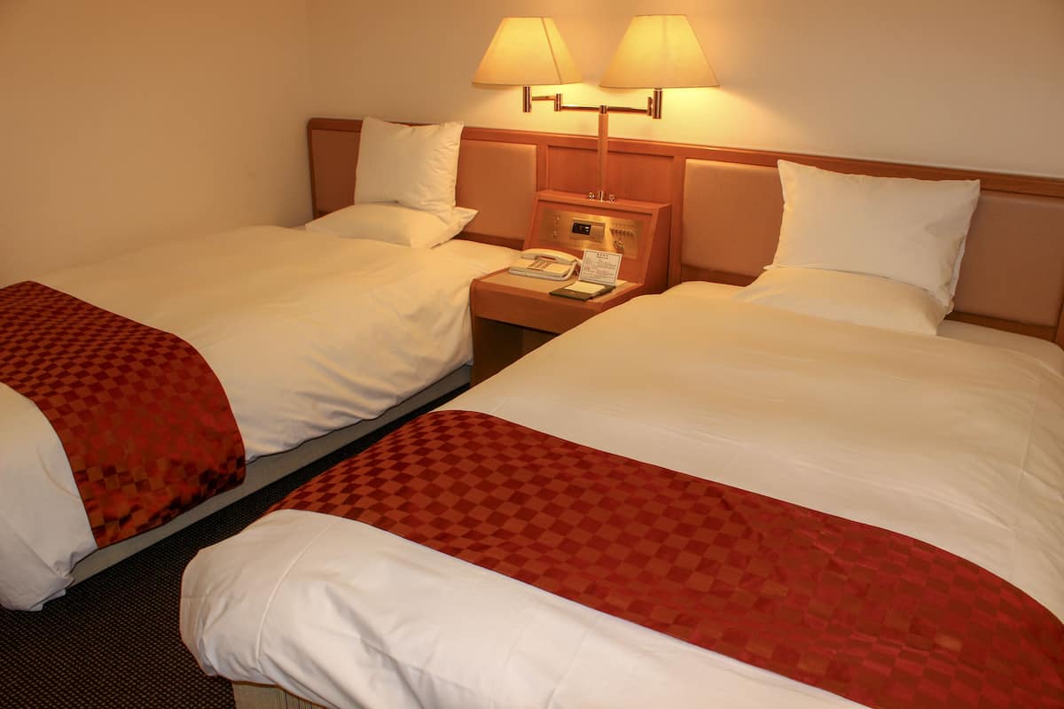 Habitación de hotel con dos camas, cada una con colcha a cuadros rojos y ropa de cama blanca. Hay dos mesitas de noche con lámparas y un pequeño reloj.
