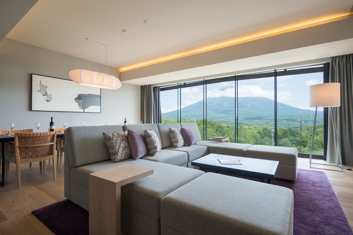 A modern hotel room with a large window offering a mountain view, a sofa set, a dining area, and a minimalist decor style.