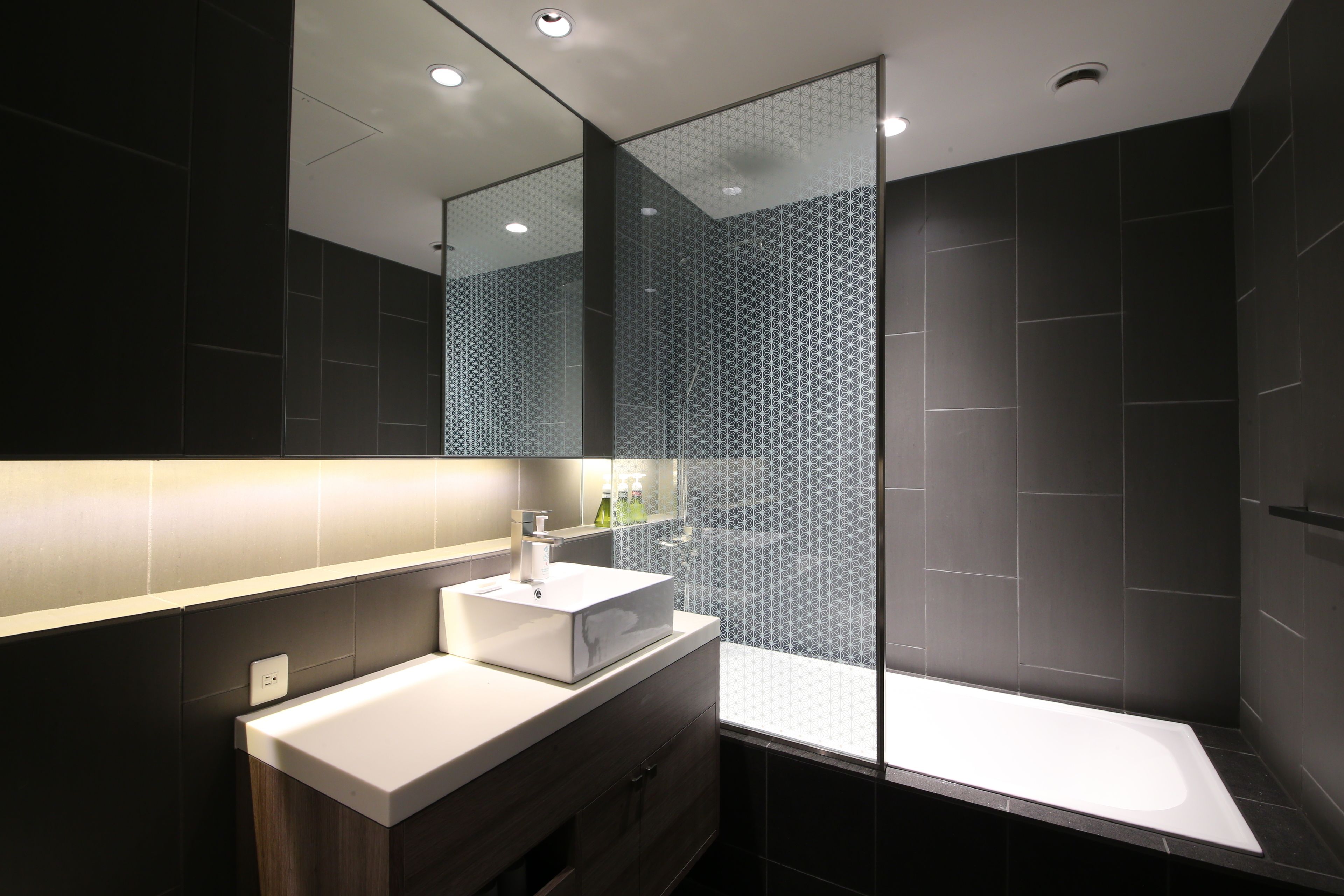 Un baño moderno con cabina de ducha de vidrio, un lavabo blanco y una pared revestida de baldosas oscuras.