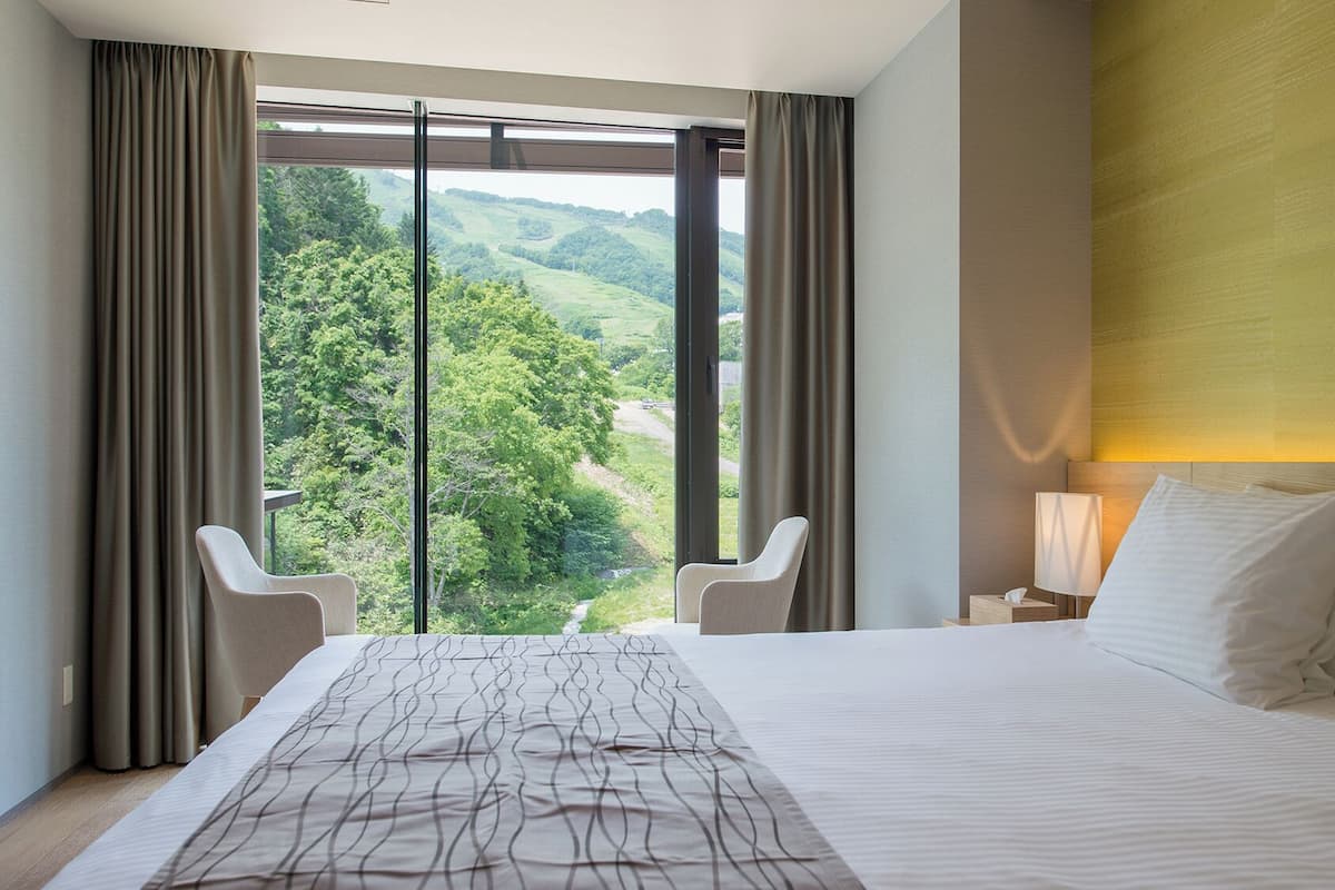 A hotel room with a large bed, a nightstand, a lamp, and a view of a green landscape through large windows.