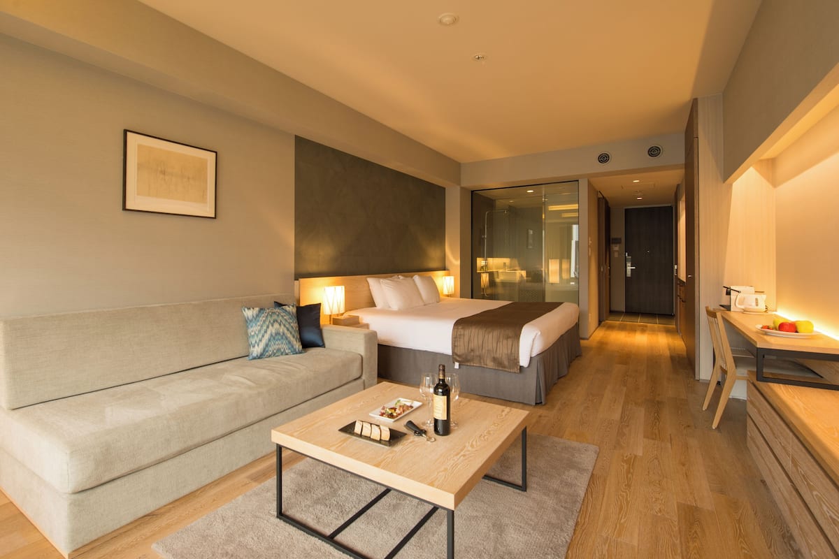 A modern hotel room with a bed, a sofa, a dining table, and a small kitchen area.