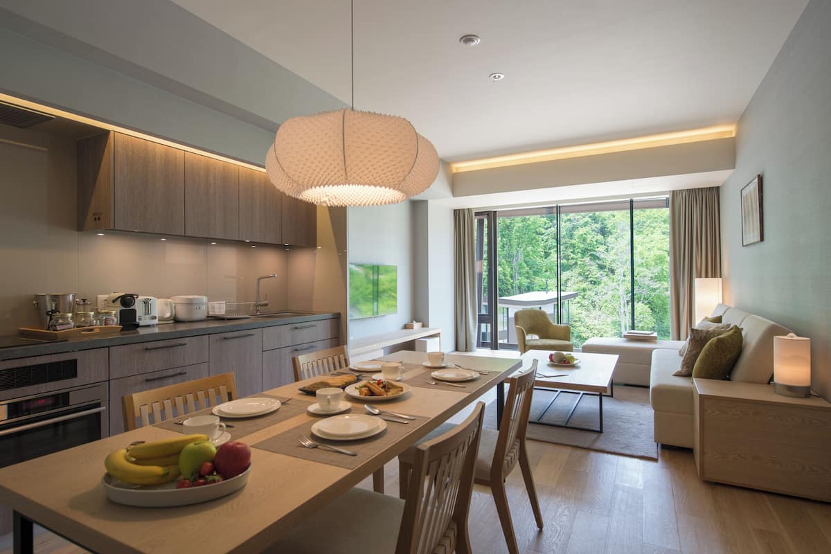 A modern kitchen with wooden cabinets, a dining table set for four, and a large window offering a view of greenery.