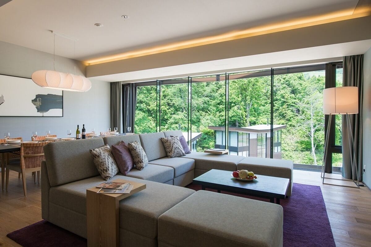 A modern living room with a sectional sofa, a dining area with a table and chairs, and large windows offering a view of greenery.