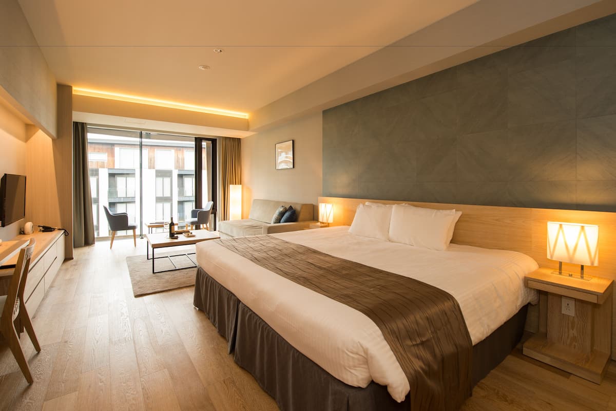 A modern hotel room with a large bed, bedside tables, a desk with a chair, a sofa, and a view of the city through large windows.