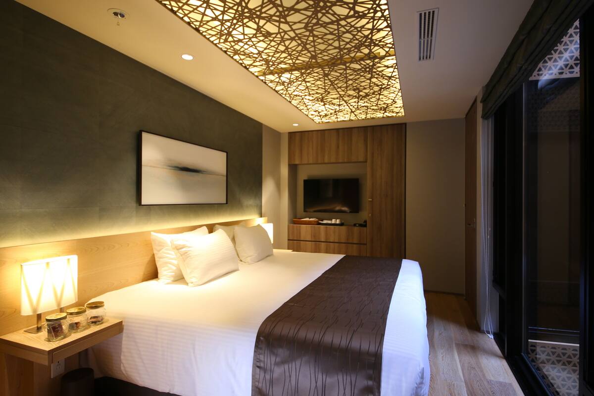 A modern hotel room with a large bed, a flat-screen TV, a wooden cabinet, and a decorative ceiling light fixture.