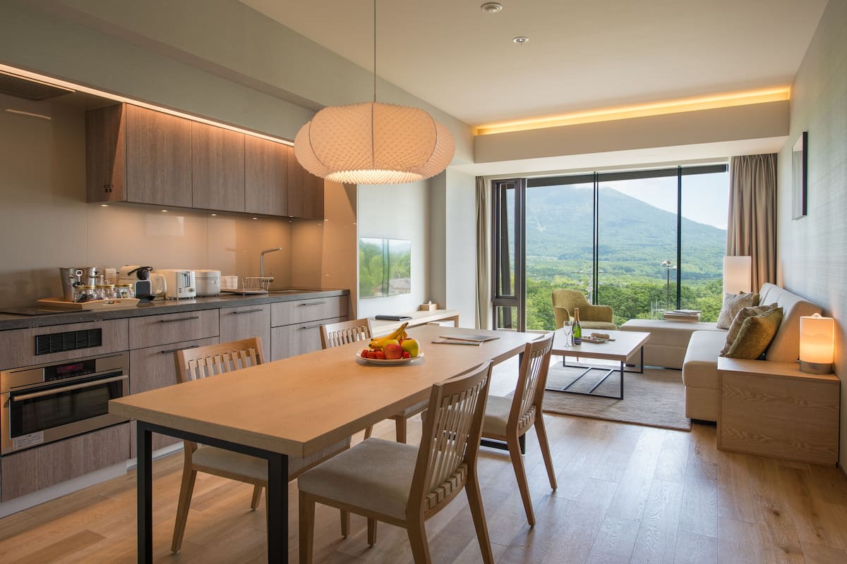 A modern kitchen with wooden cabinets, a dining table with chairs, and a large window offering a view of a mountain.