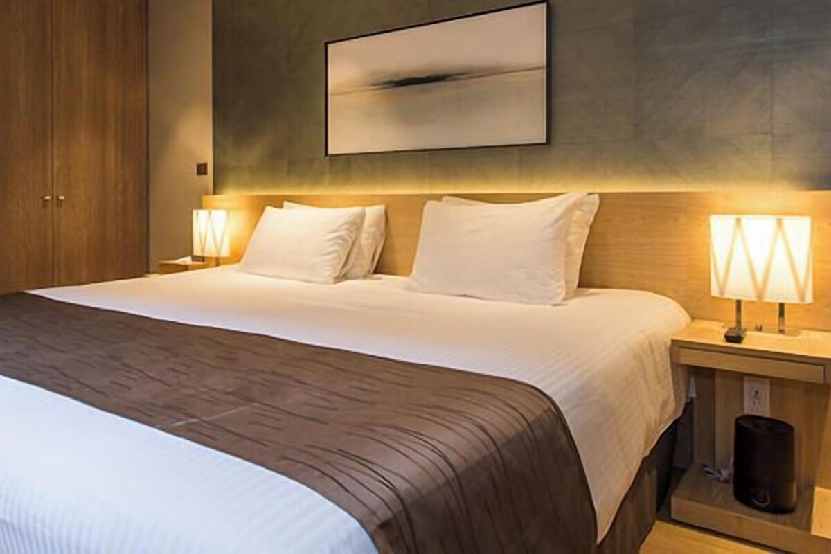 A hotel room with a large bed, two bedside lamps, a wooden headboard, a wall-mounted picture, and a small table with a trash bin.