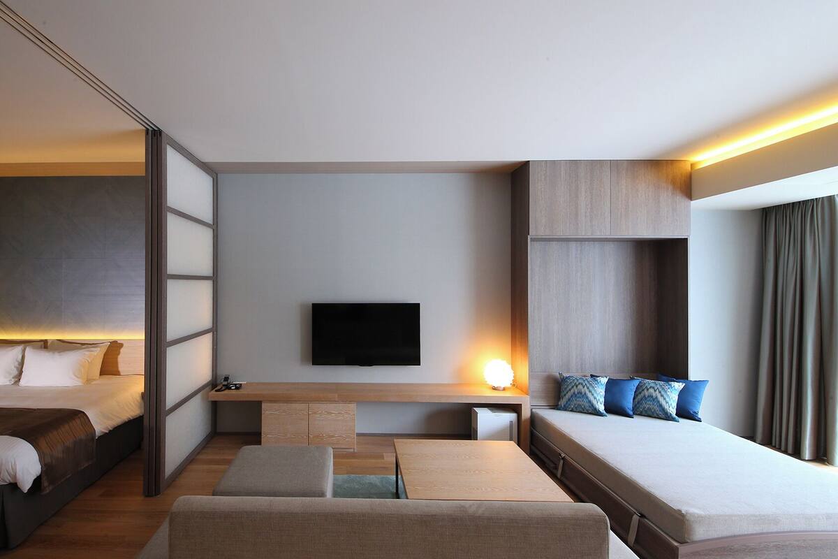 A modern hotel room with two beds, a flat-screen TV, a small table, and a seating area.