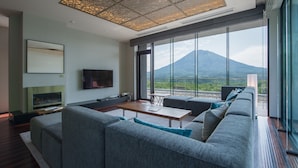 A modern living room with a large sofa, a flat-screen TV, and a dining table. A large window offers a view of a mountain.