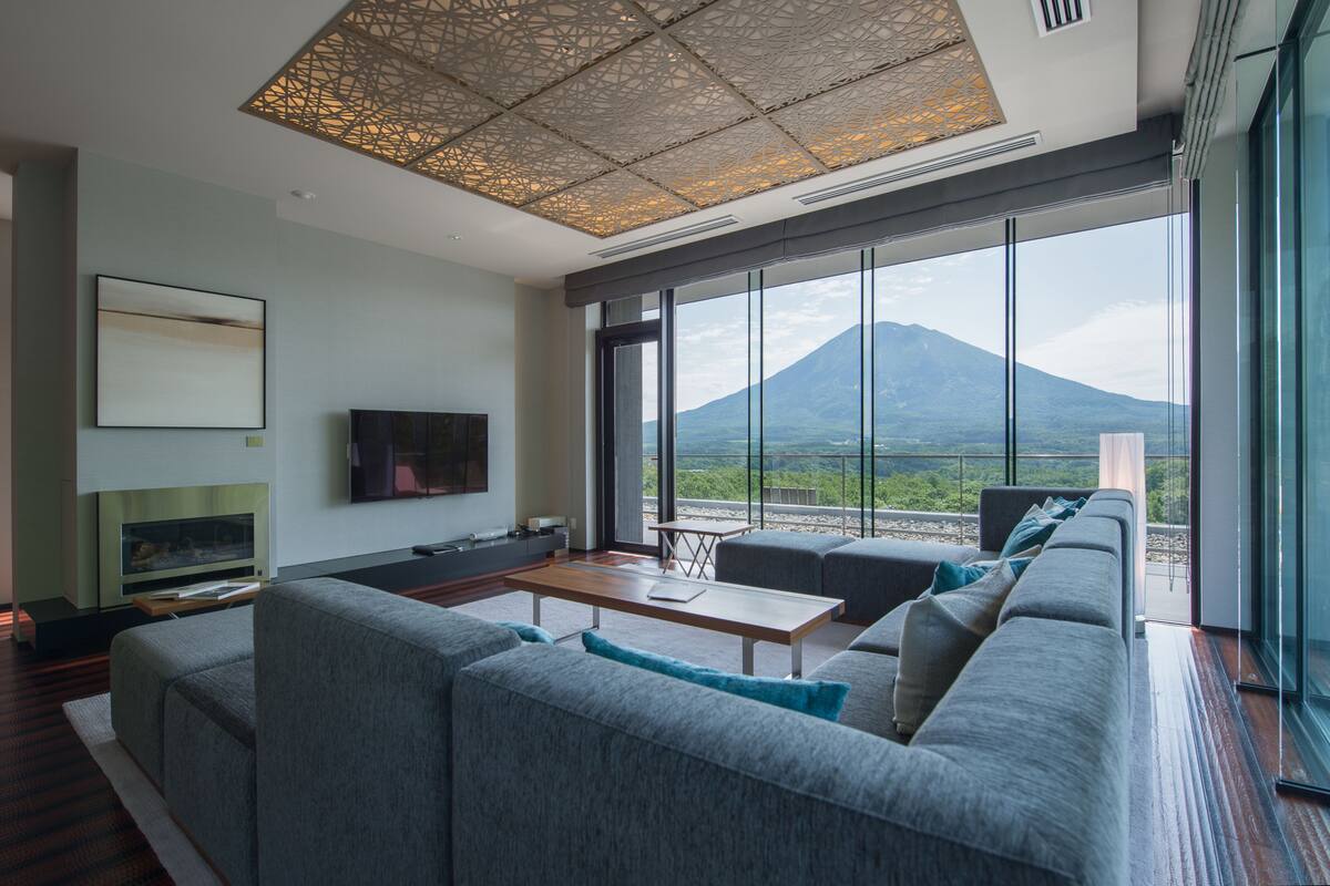A modern living room with a large sofa, a flat-screen TV, and a dining table. A large window offers a view of a mountain.