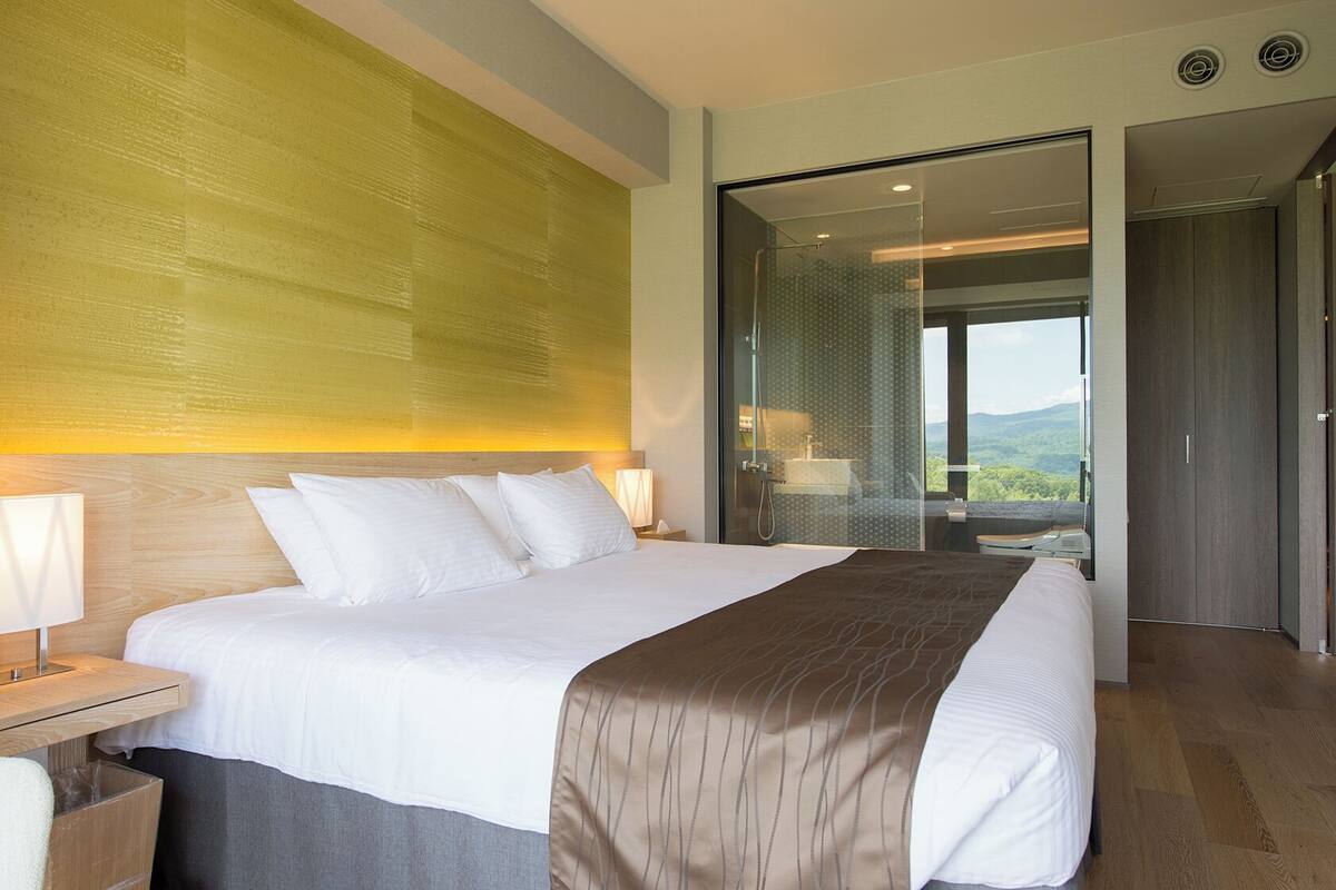 A hotel room with a large bed, a nightstand, a lamp, a bathroom with a shower, and a view of the outdoors.