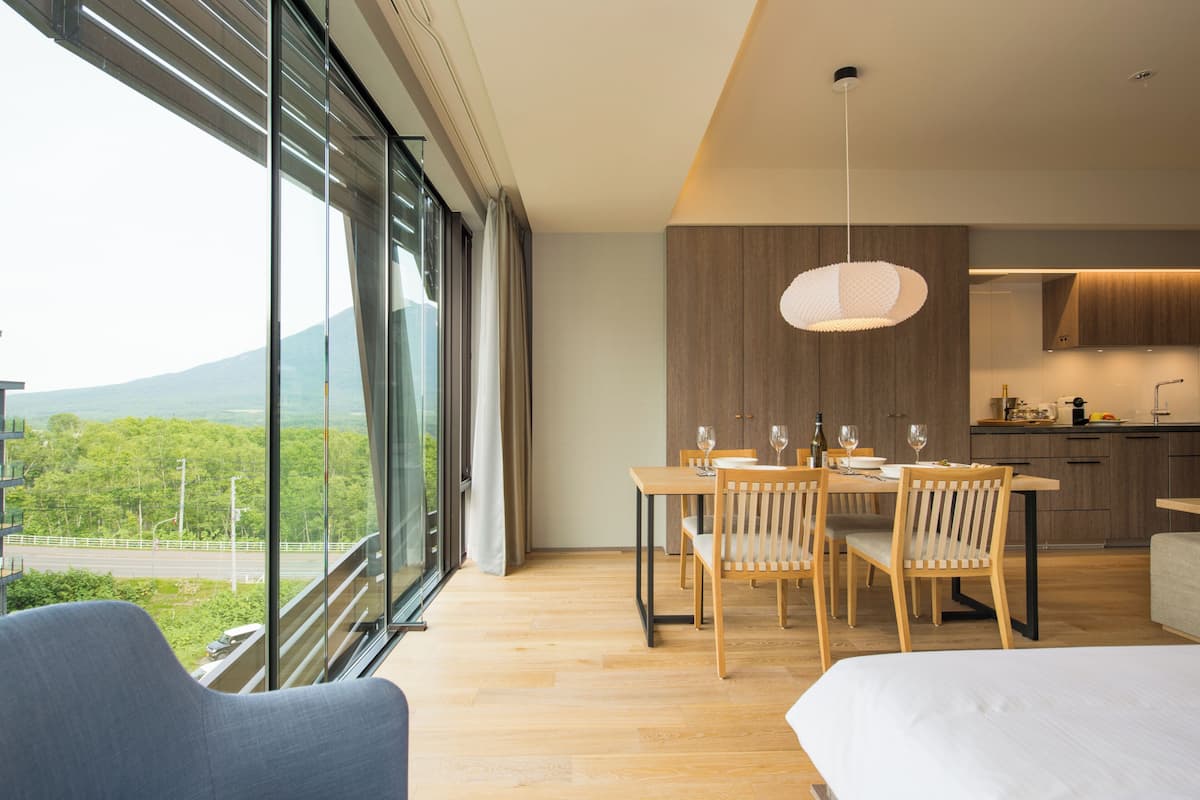 A modern hotel room with a large window, a dining area with wooden furniture, and a kitchen area.
