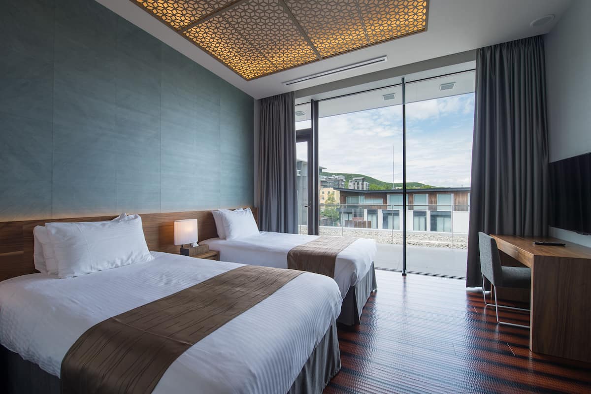 A hotel room with two beds, a desk, and a large window offering a view of the city.