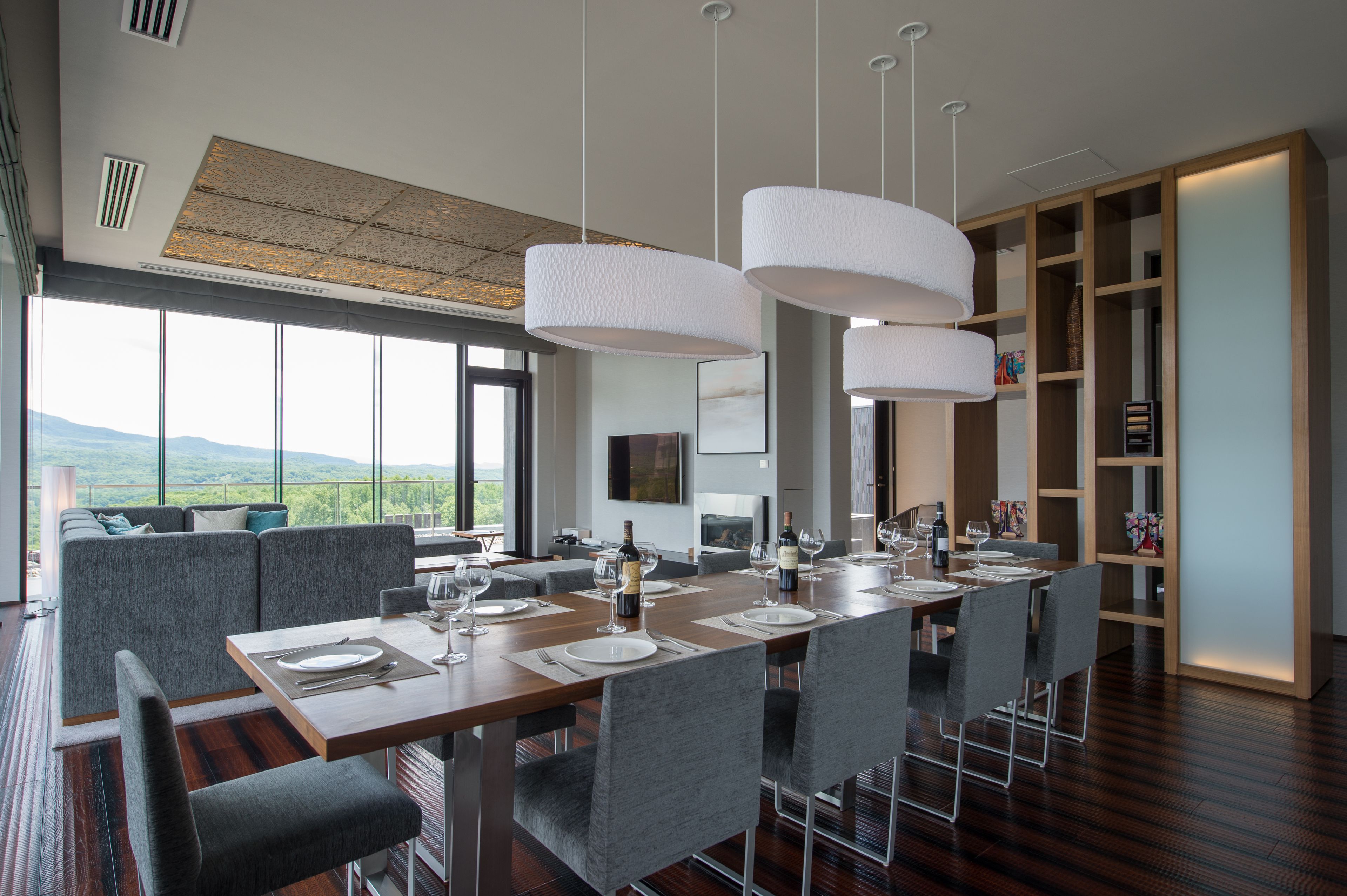 Un moderno comedor con una larga mesa de madera dispuesta para seis personas, rodeada de sillas grises tapizadas. Hay lámparas colgantes blancas sobre la mesa, y un gran ventanal ofrece vistas al exterior.