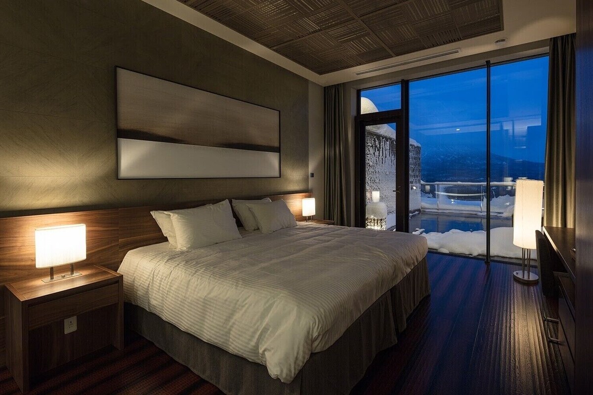 A modern hotel room with a large bed, bedside lamps, a large window with a view of a snowy landscape, and a ceiling with a textured pattern.