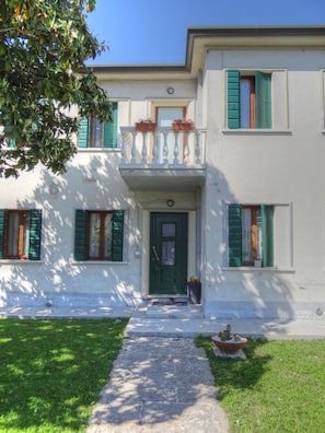 A two-story building with a green door, white walls, and green shutters. There is a small balcony with potted plants and a flower box. A concrete pathway leads to the entrance. - B&B Villa Griselda (Quinto di Treviso)