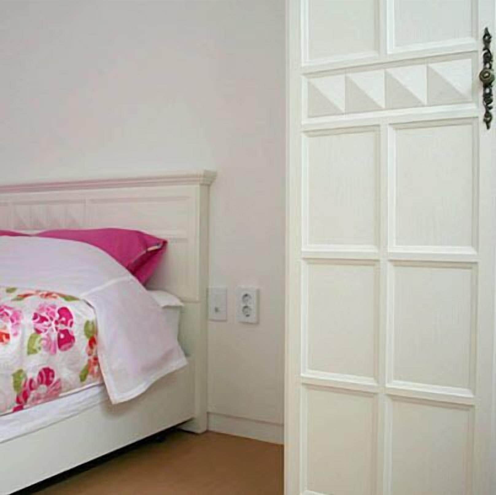 Un lit blanc, recouvert d’une couvre-lit à motifs floraux, avec un oreiller rose, dans une pièce aux murs et à la porte blancs.