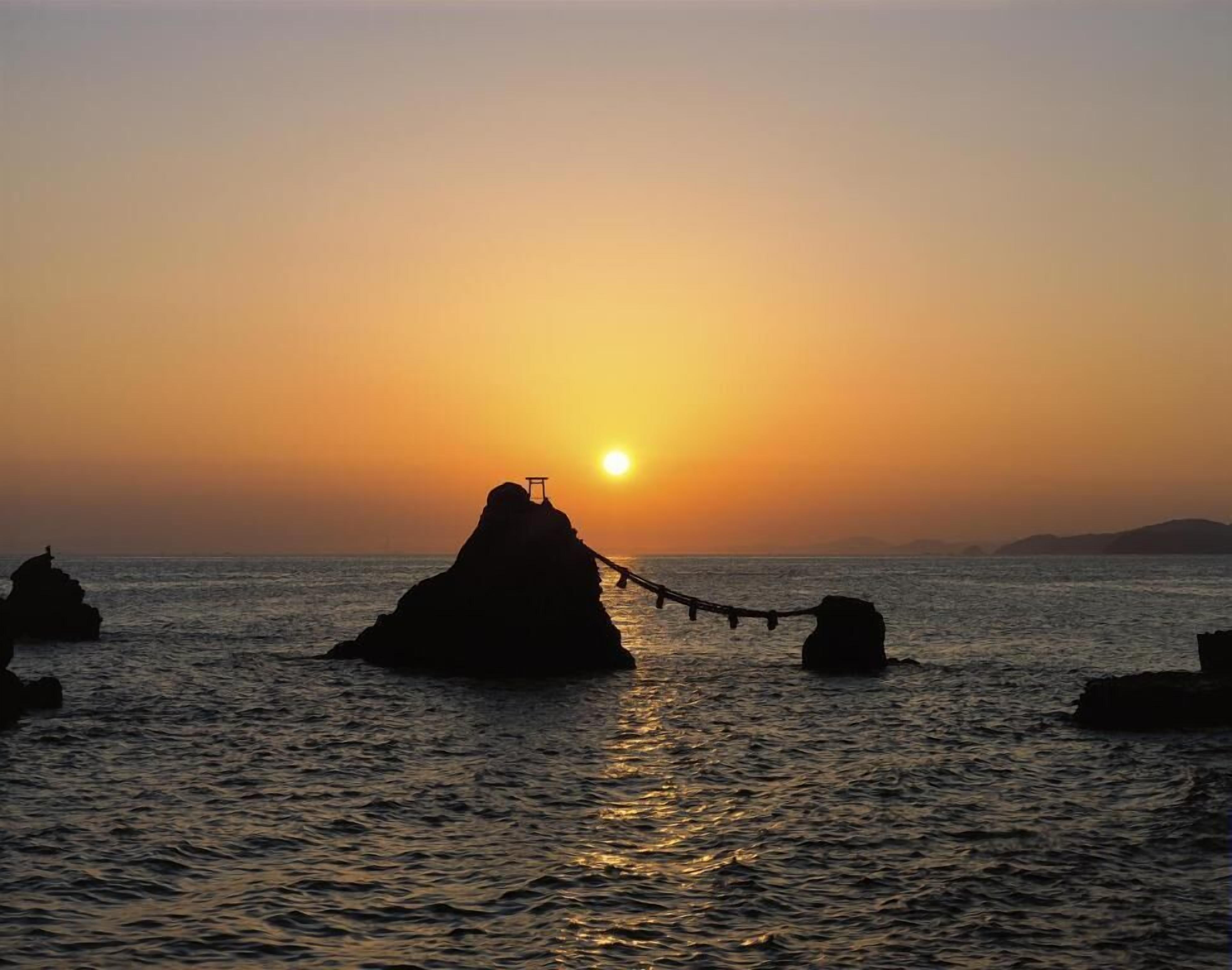 A sunset over a calm sea with a rock formation in the center and a rope bridge leading to it.