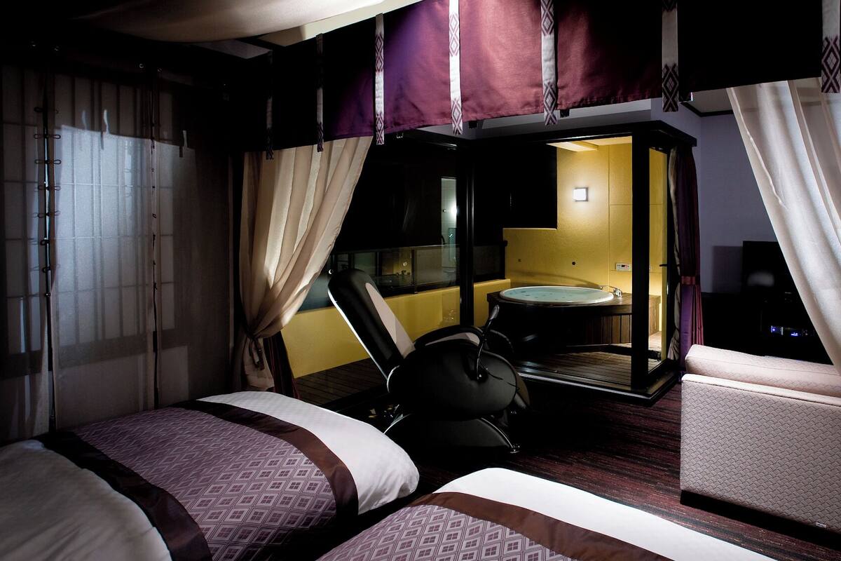 Una habitación de hotel con dos camas, una mesita, una silla y un baño visible a través de una puerta abierta.