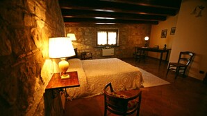 Un dormitorio acogedor con pared de piedra, cama con colcha blanca, mesita de noche de madera con lámpara y una silla.