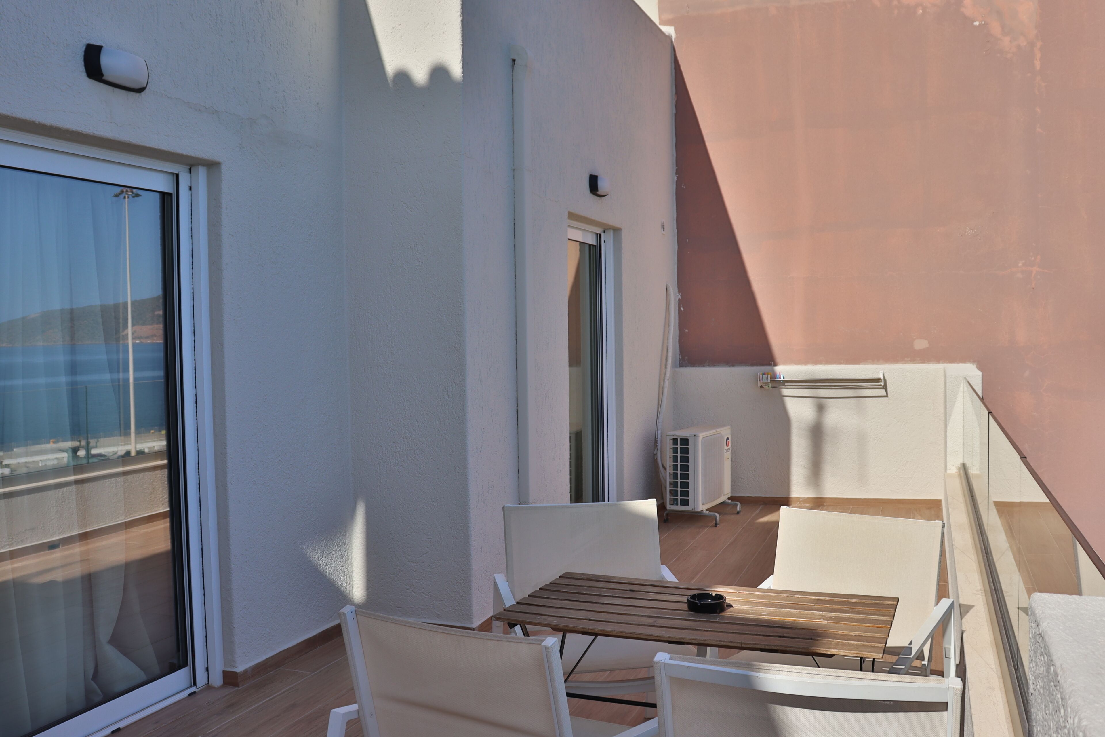Un balcone con tavolo in legno e sedie bianche, una porta scorrevole in vetro e un piccolo condizionatore d'aria.