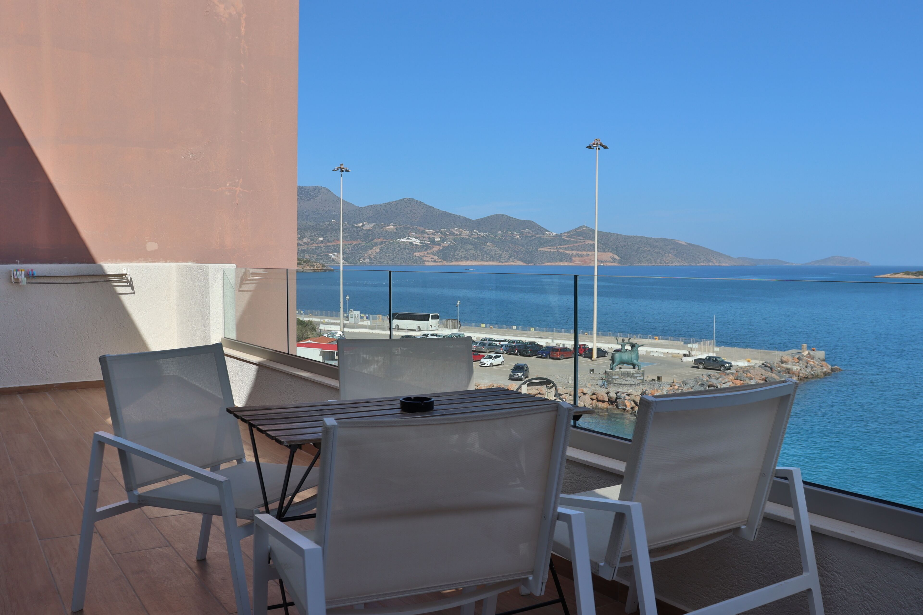 A balcony with a table and chairs, overlooking a coastal area with a beach, parked cars, and mountains in the background.