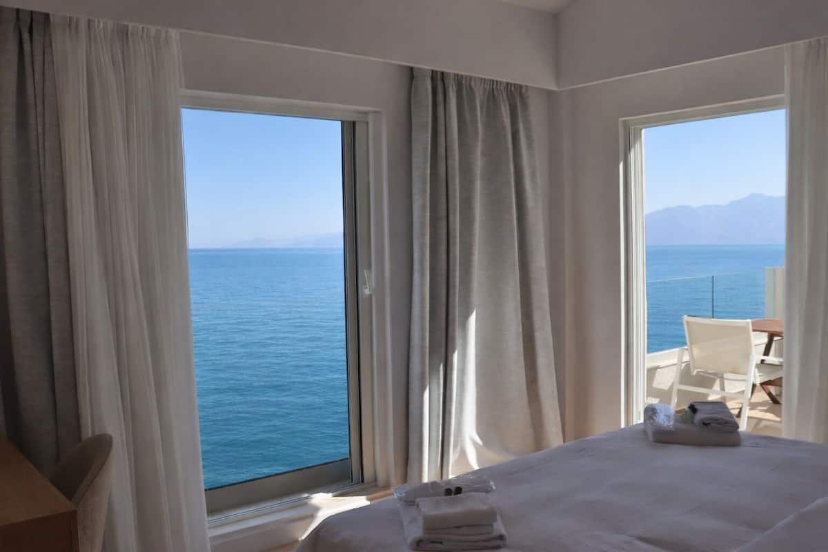 A hotel room with a large window offering a sea view, a bed with neatly folded towels, and a chair by the window.