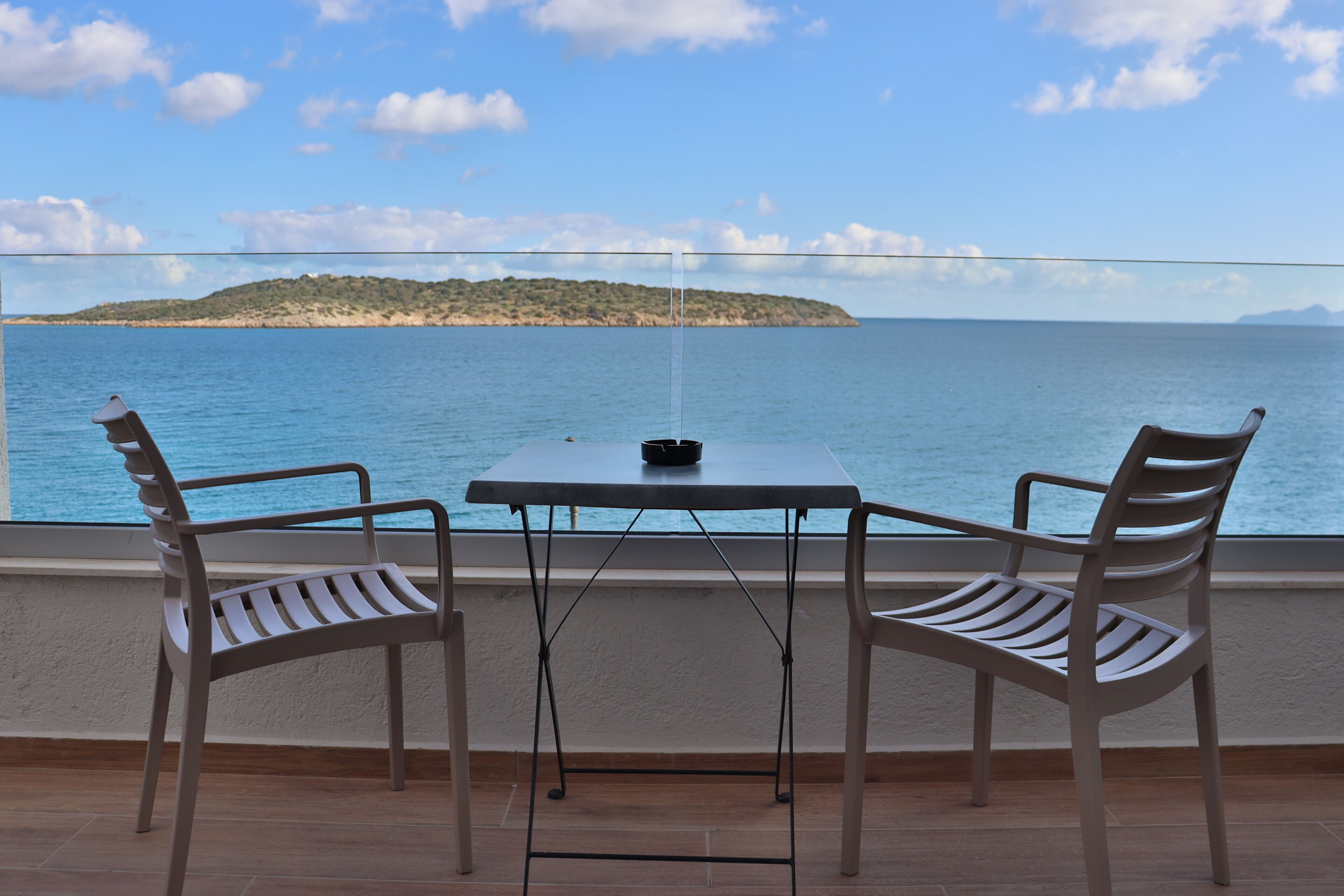 A balcony with two chairs and a table, overlooking a calm sea and a distant island.