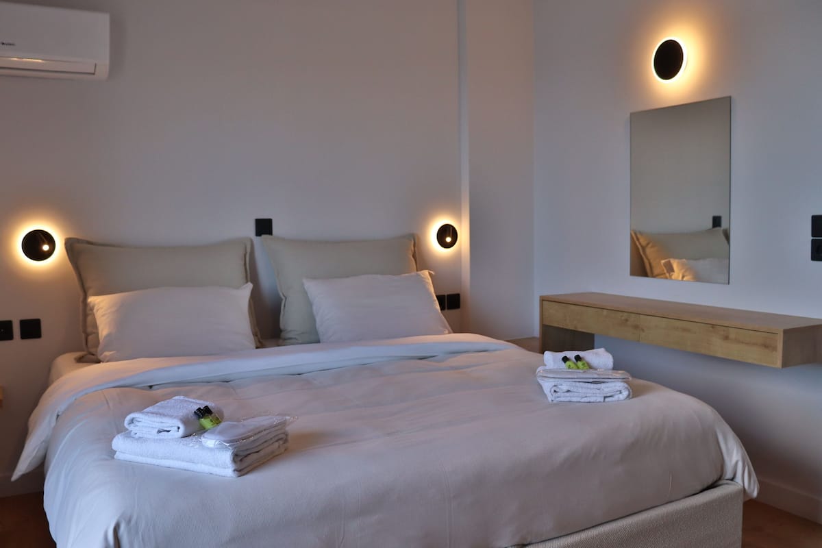 A modern hotel room with a neatly made bed, a wooden bedside table with towels, and a wall-mounted mirror.