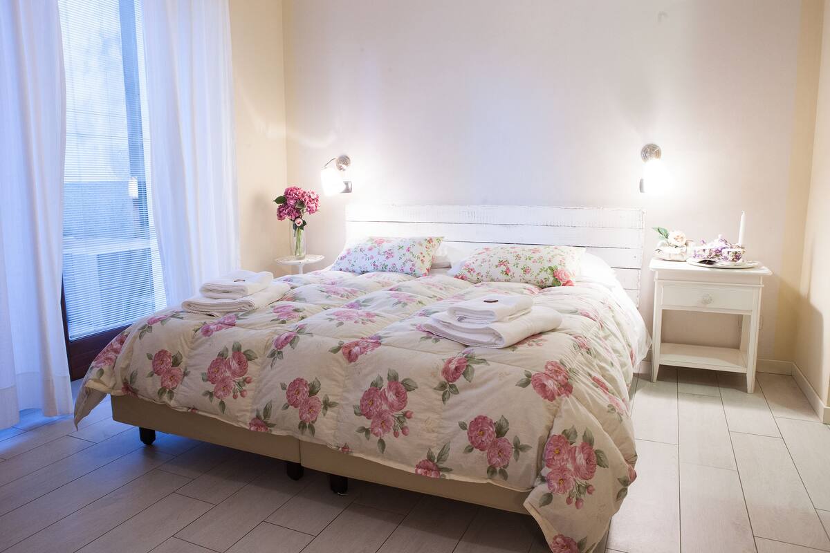 A neatly made bed with floral bedding, a bedside table with a lamp, and a vase with flowers.