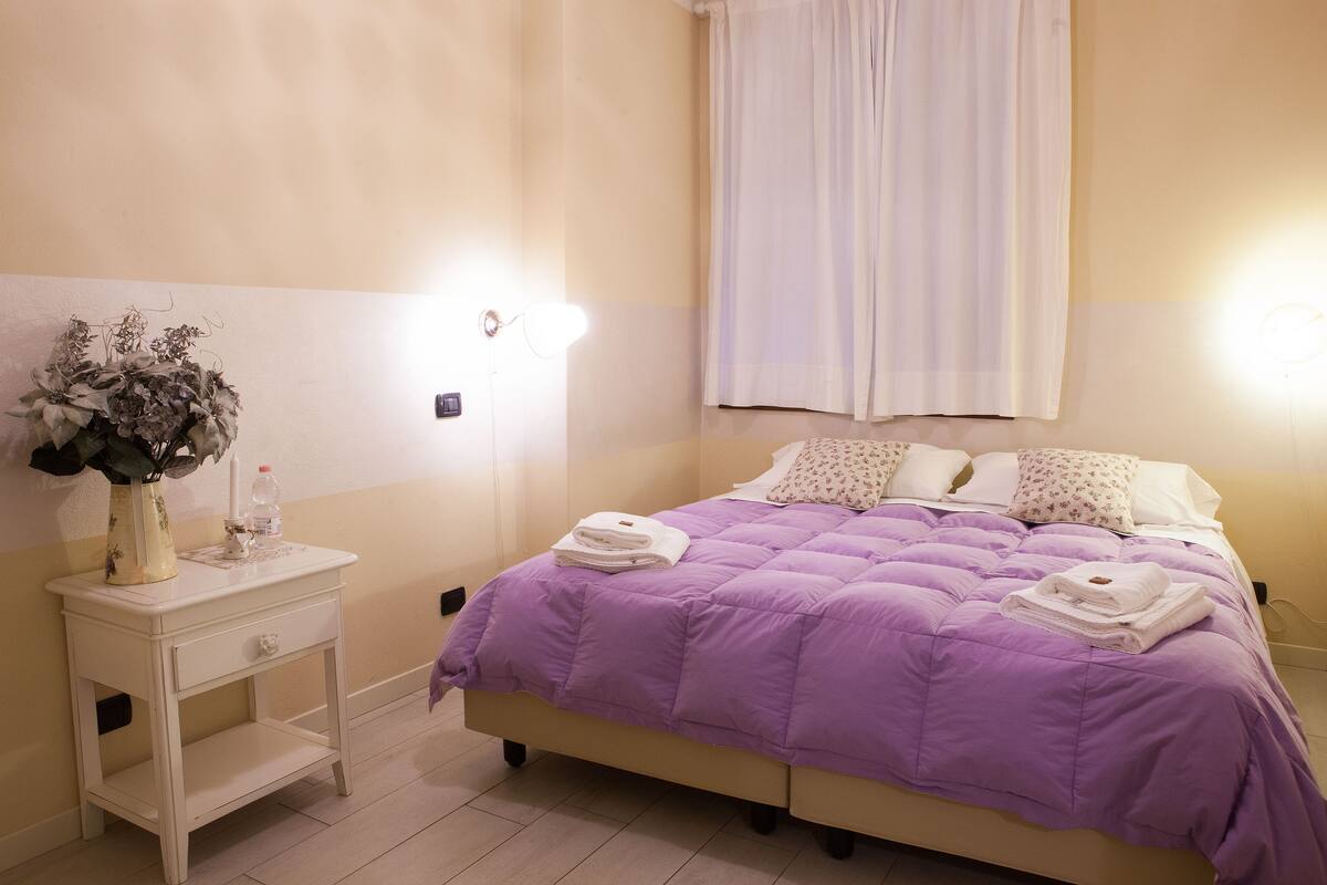 A neatly made bed with a purple bedspread, a bedside table with a vase and a bottle, and a wall-mounted light.