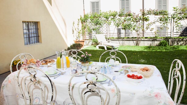 A table set for breakfast with a white tablecloth, metal chairs, and a view of a building with shutters and greenery in the background. - Botton d'Oro Pelabrocco (Bergamo)