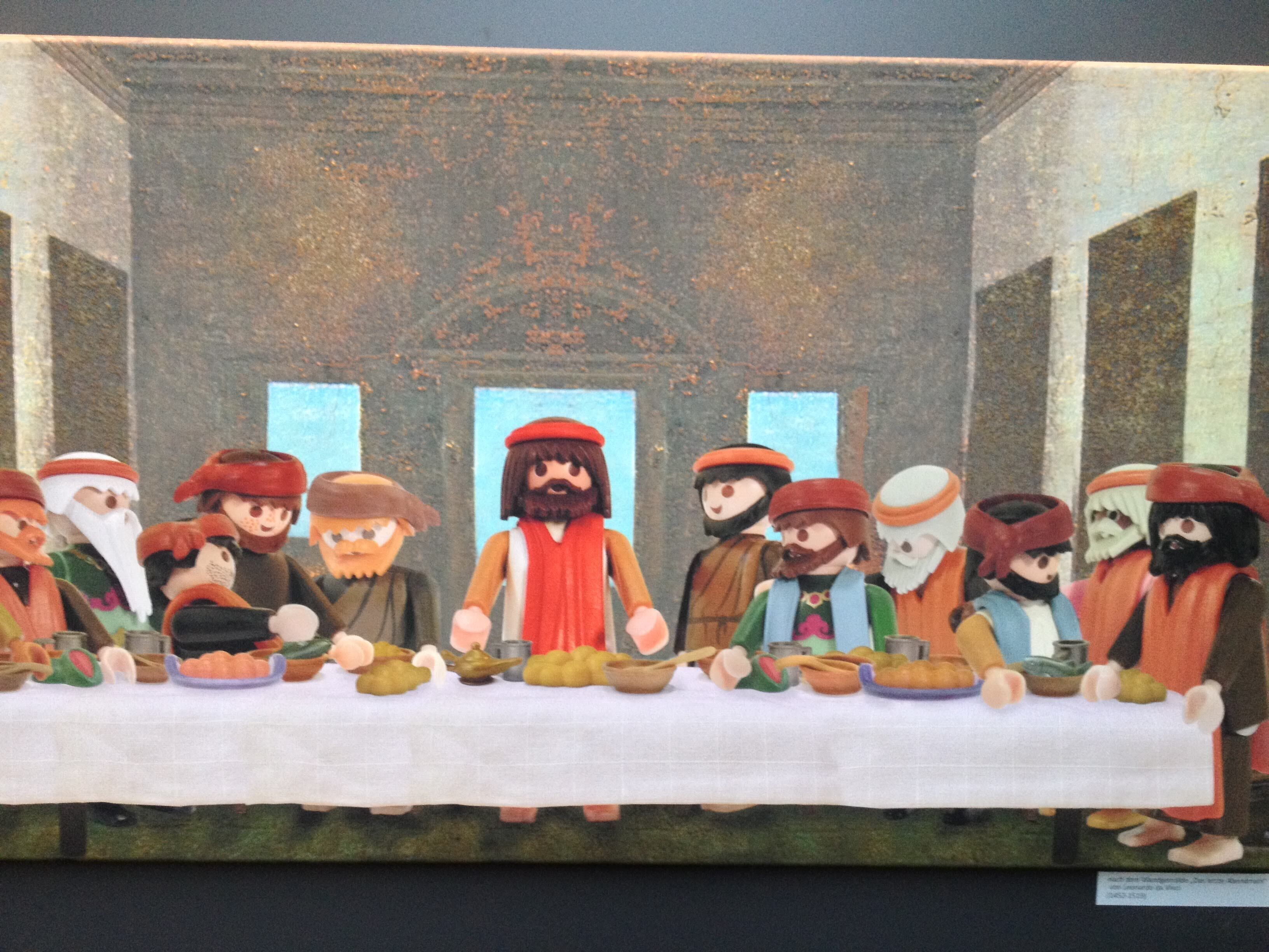 Una pintura de un grupo de personas sentadas a una mesa con comida, con un edificio con ventanas de fondo.