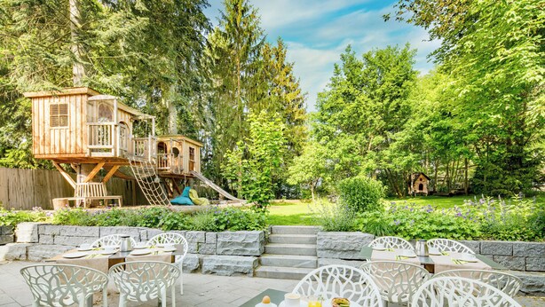 An outdoor dining area with white chairs and tables, surrounded by a lush garden and a wooden playhouse. - Hotel Knorz (Zirndorf)