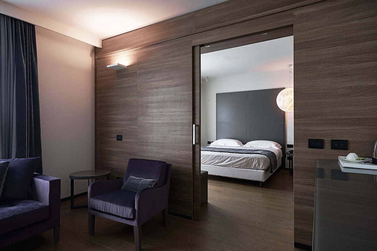 A modern hotel room with a wooden accent wall, a large mirror, a bed with a grey headboard, a small round table, and a dark blue armchair.