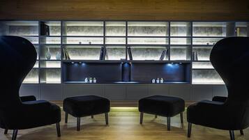 Una habitación moderna con suelo de madera, un sofá oscuro, dos otomanas y una estantería con repisas iluminadas.