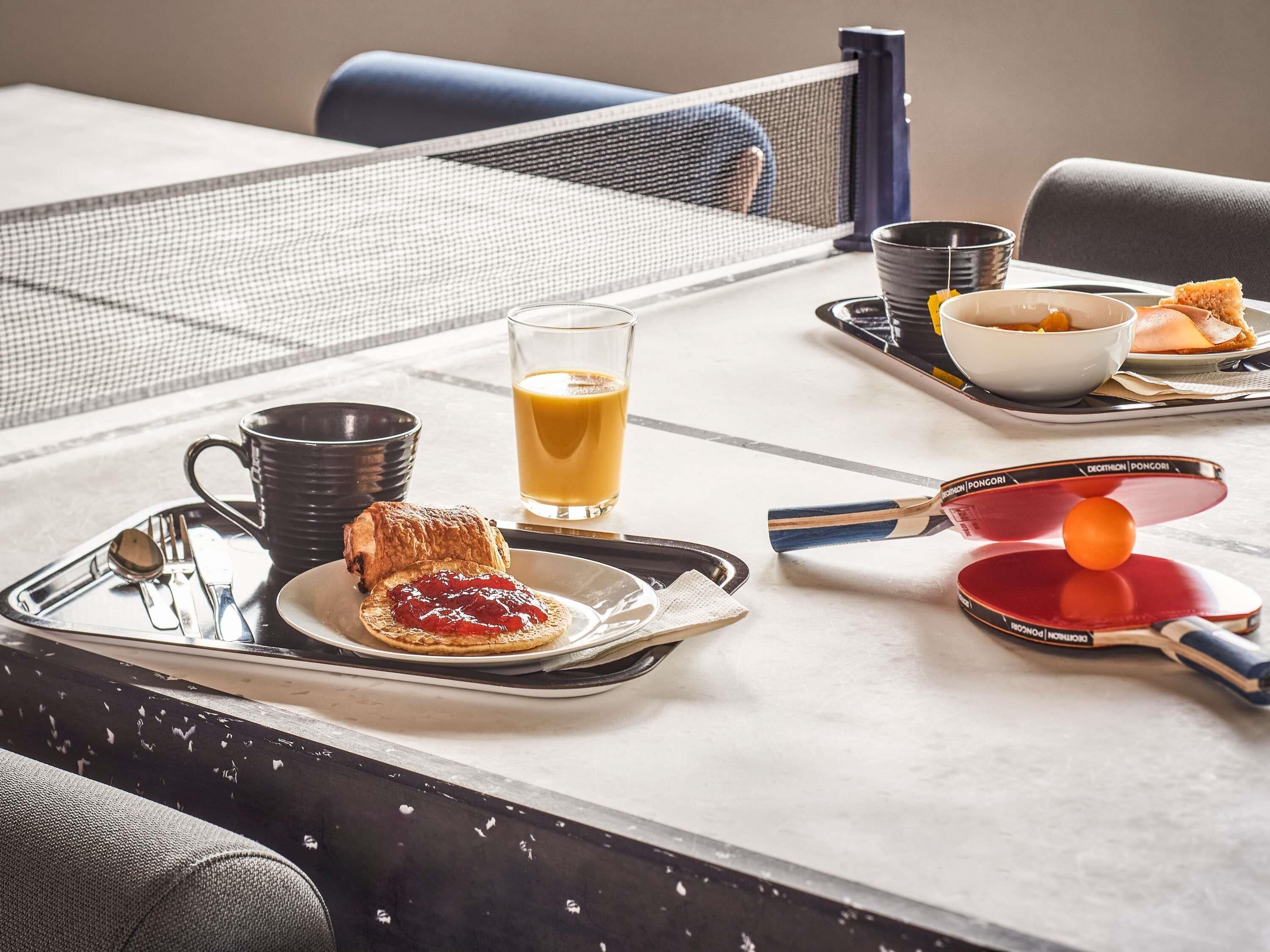 Bandeja de desayuno con tostadas, mermelada, un vaso de zumo de naranja, una taza y una paleta de ping pong sobre una mesa.