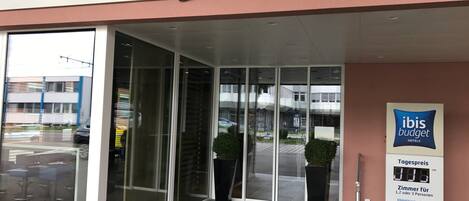The exterior of an ibis budget hotel with a glass entrance, signage indicating the price per room, and a view of the street outside.