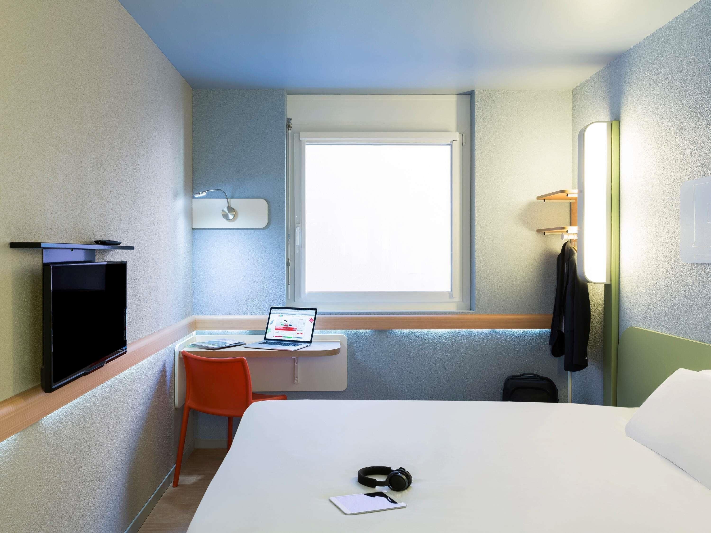 Une chambre d’hôtel comprenant un lit, un bureau avec un ordinateur portable, une chaise, une télévision, une fenêtre et un porte-manteau.