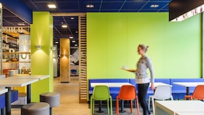 Ein modernes Café mit einer hellgrünen Wand, Holzelementen und bunten Stühlen.