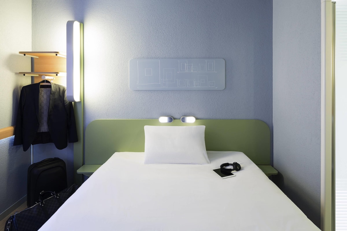 A hotel room with a bed, a wall-mounted light, a headboard with two lights, a wall map, a hanging coat, and a suitcase.