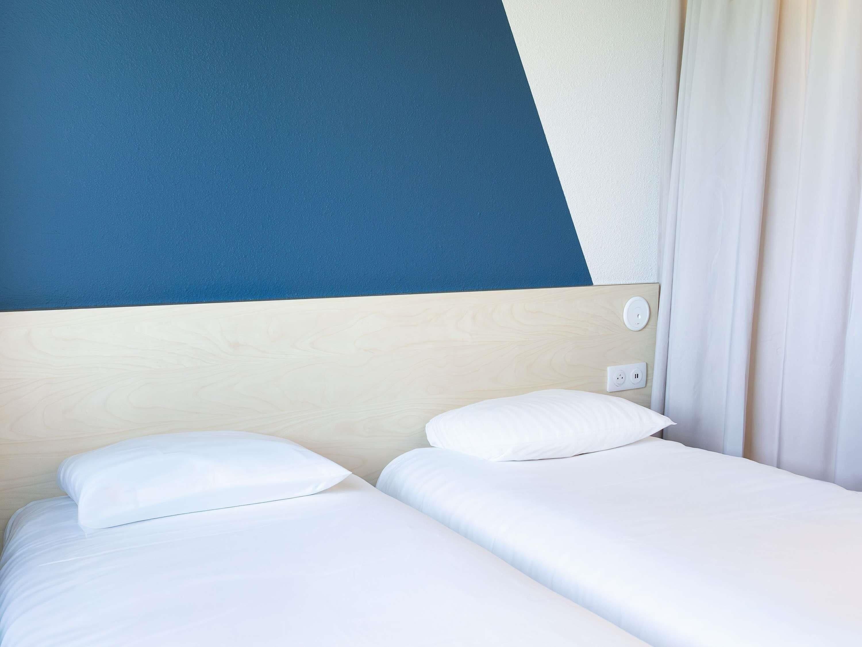 A hotel room with a bed, a nightstand, a power outlet, and a window with curtains.