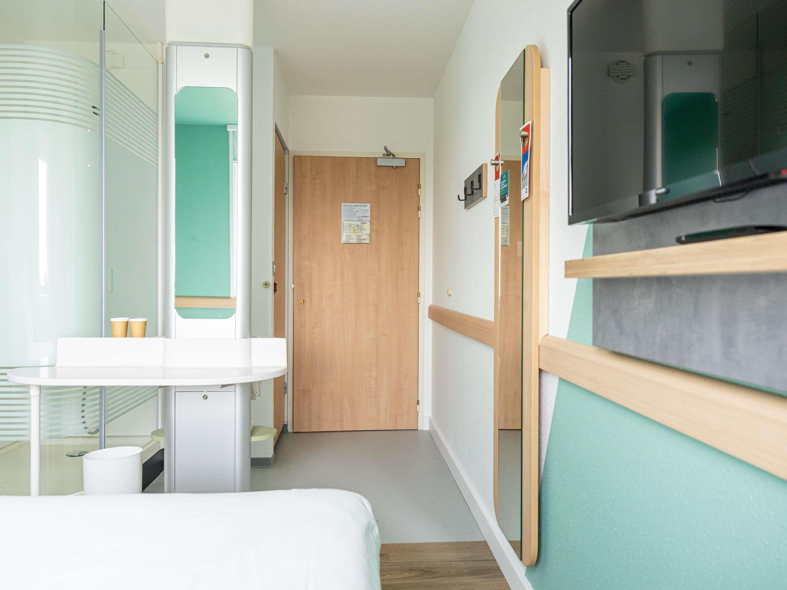 A hotel room with a bed, a small desk, a mirror, a wooden door, and a wall-mounted TV.