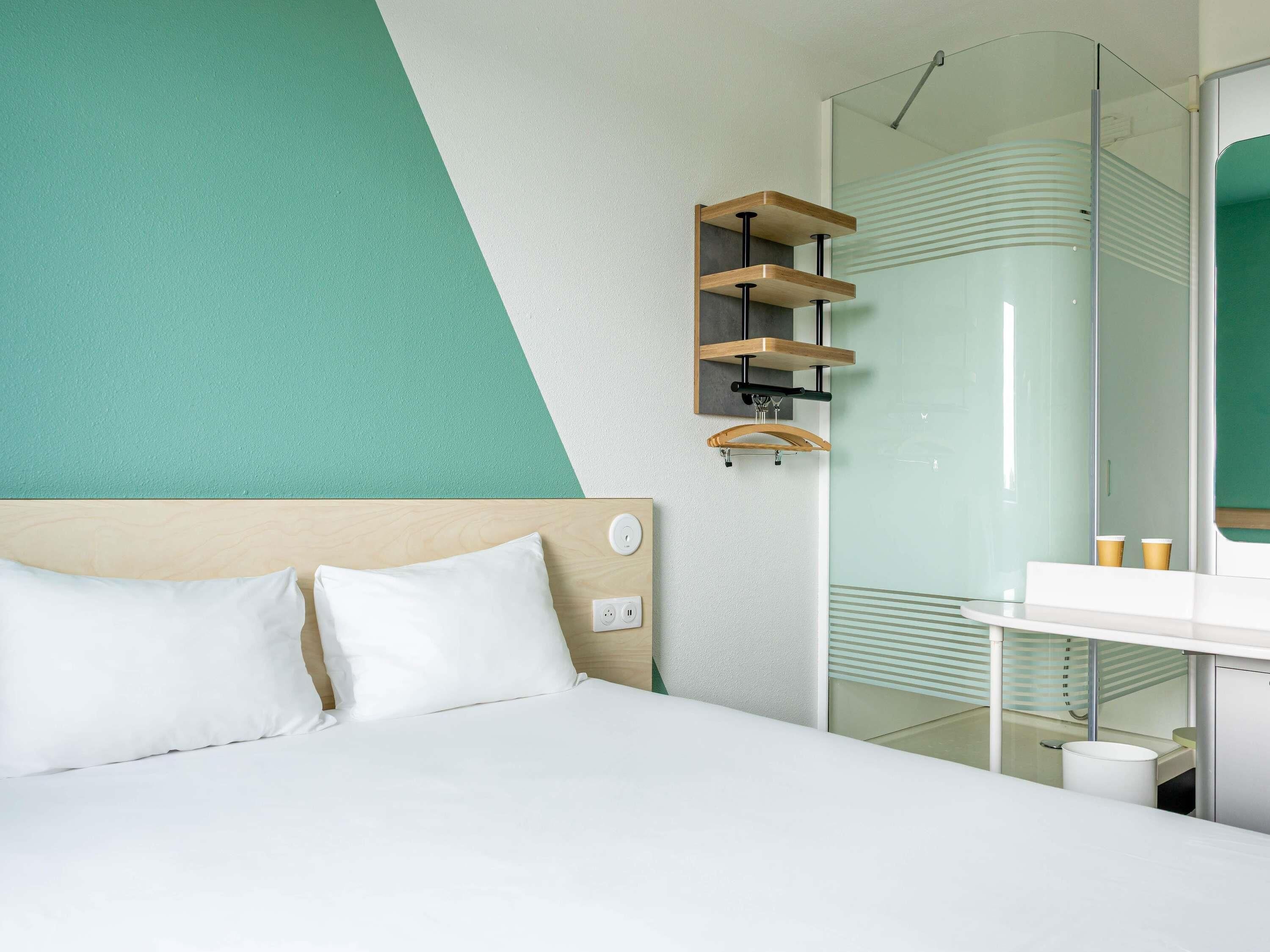 A modern hotel room with a bed, a glass shower, and a wooden shelf unit.