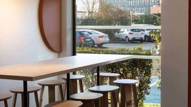 Un interior moderno con una mesa larga y varios taburetes, un ventanal con vistas a coches aparcados y edificios.