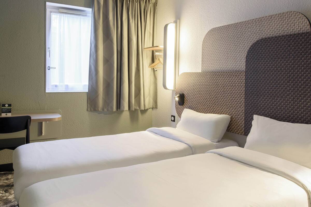 A hotel room with two beds, a desk, and a window with curtains.