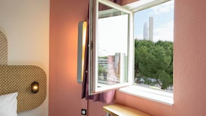 A room with a window, a bed, a wall-mounted light, and a view of a building and trees outside.