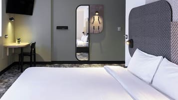 Et hotelværelse med seng, skrivebord, stol, tv ophængt på væggen og et spejl der reflekterer rummet.